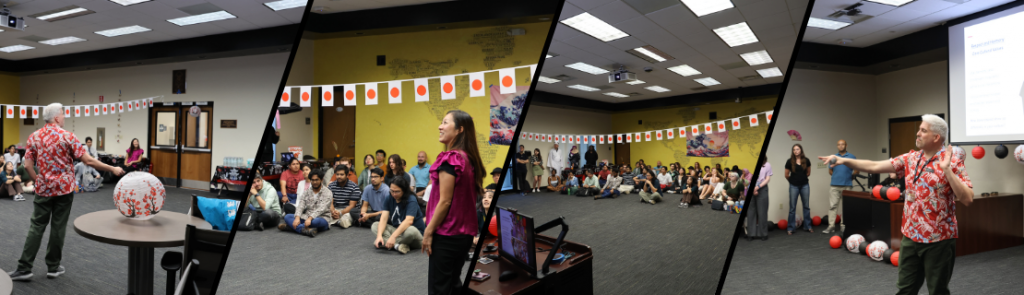 A cultural event in a large room decorated with Japanese flags and lanterns. People are seated on the floor and chairs, listening to presenters. A person in a floral shirt holds a microphone and gestures toward the audience, while another stands near a table with props.