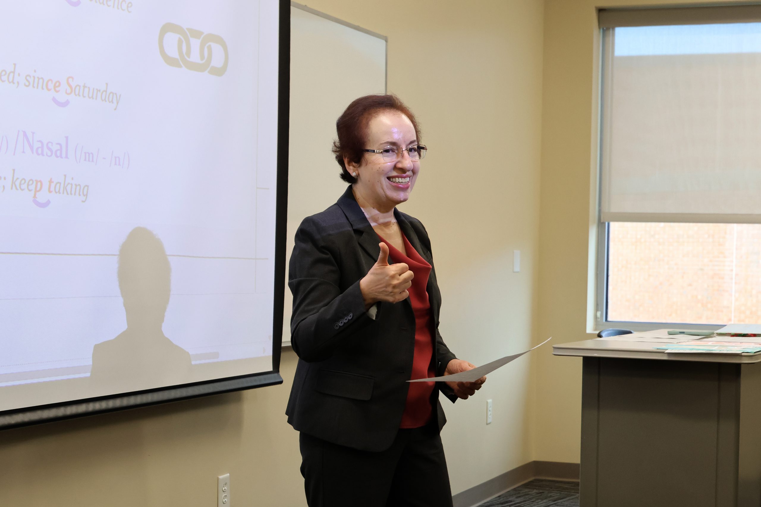 A person in business attire stands at the front of a classroom giving a presentation, holding a sheet of paper in one hand and gesturing with the other. A projection screen with text is displayed beside them, and a desk with materials is visible near the window.