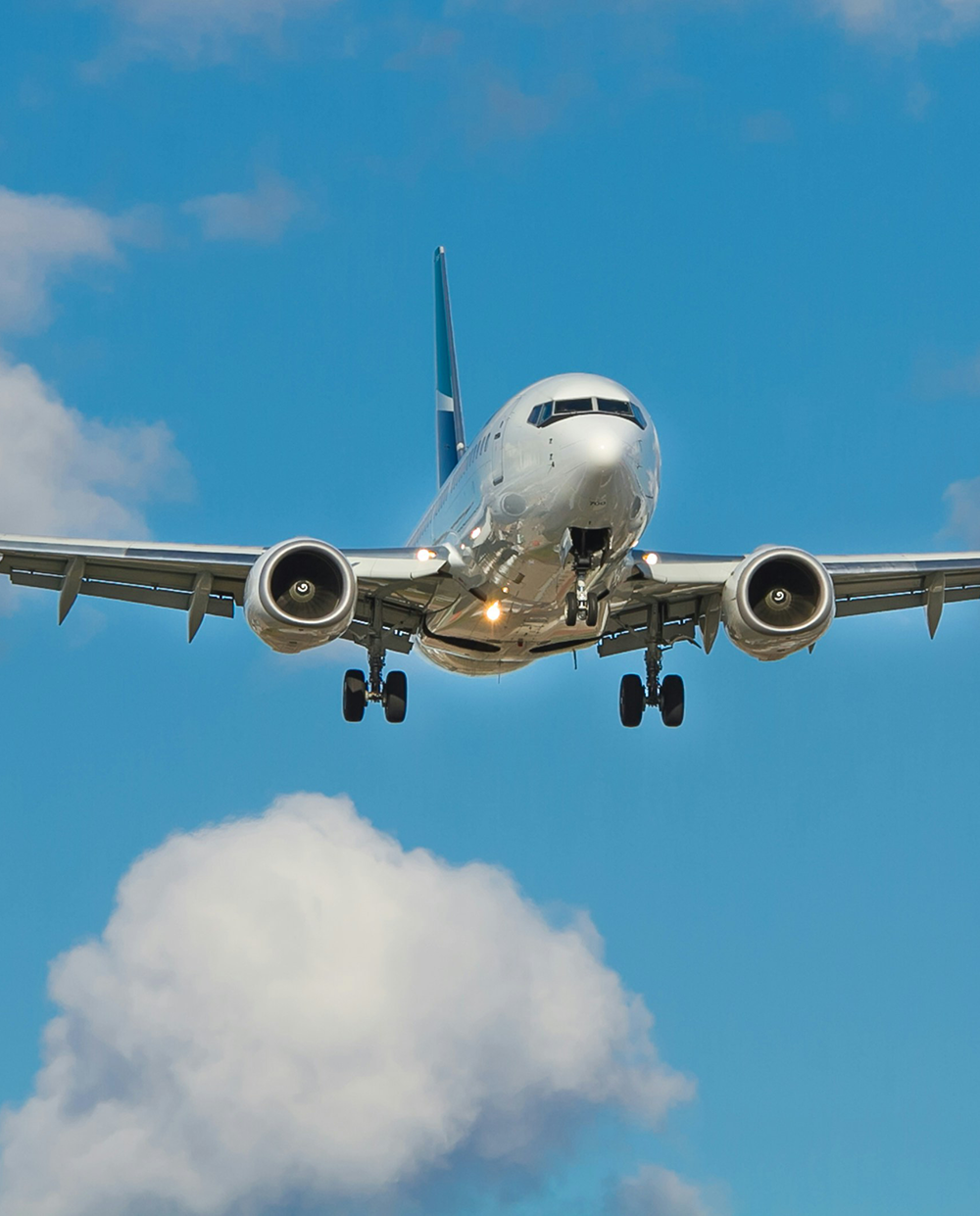 A commercial airplane approaching head‑on against a bright blue sky with scattered clouds, its landing gear extended and both engines clearly visible as it descends.